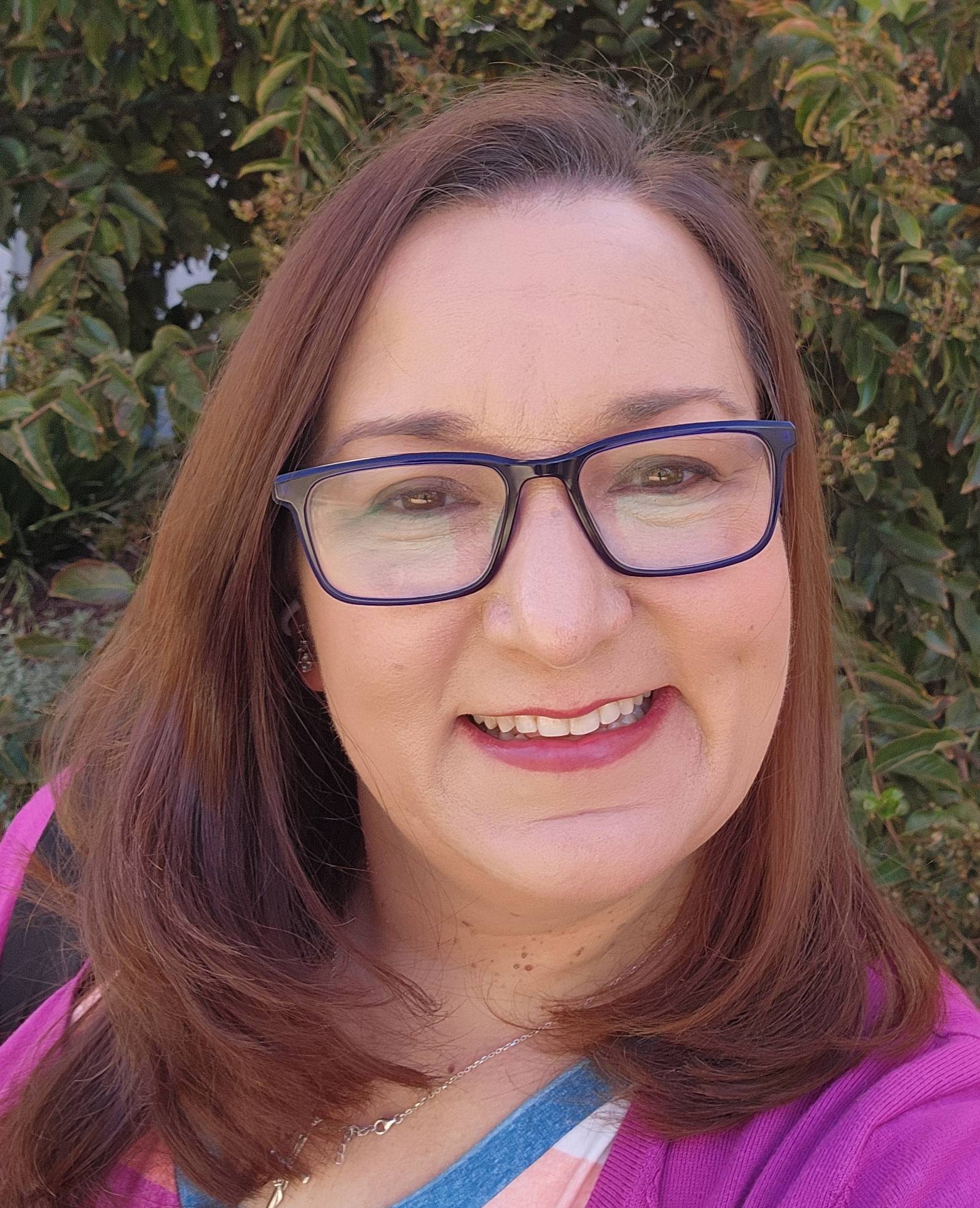 Woman's face with long brown hair, wearing blue glasses and a purple shirt