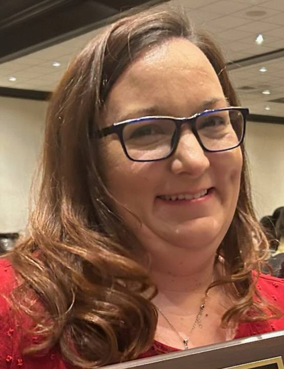 Woman's face with long brown hair, wearing blue glasses and a red shirt